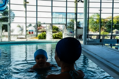 Un adulte accompagne un nourrisson dans le bassin intérieur d'un centre aquatique couvert, avec jouets gonflables colorés au-dessus de l'eau