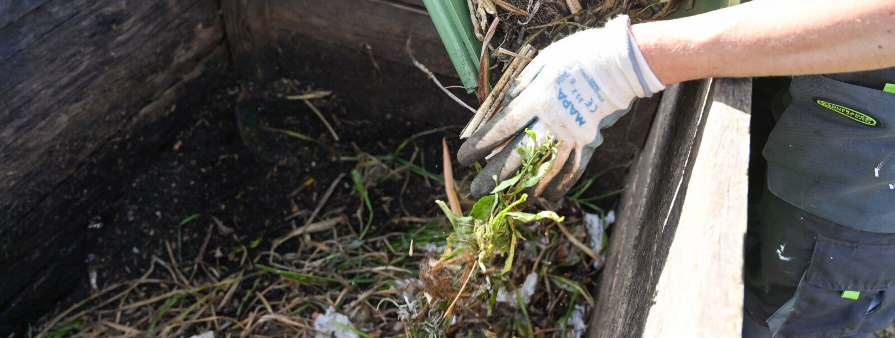 Versement de déchets verts dans un composteur en bois par une personne équipée de gants de jardinage