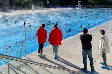 Quatre membres du staff observent depuis le bord les nageuses s'entraînant en couloirs dans le bassin extérieur.