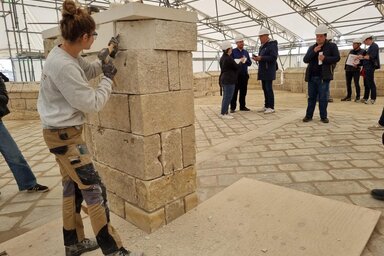 Une tailleuse de pierre façonne un pilier en tuffeau sous une tente de chantier, devant un groupe d'élus et journalistes casqués.