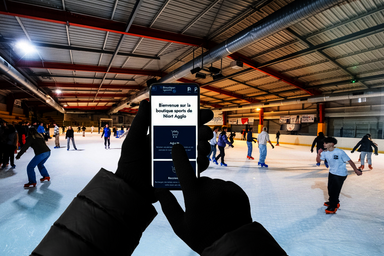 Smartphone affichant la boutique en ligne Niort Agglo, tenu devant une patinoire avec des patineurs